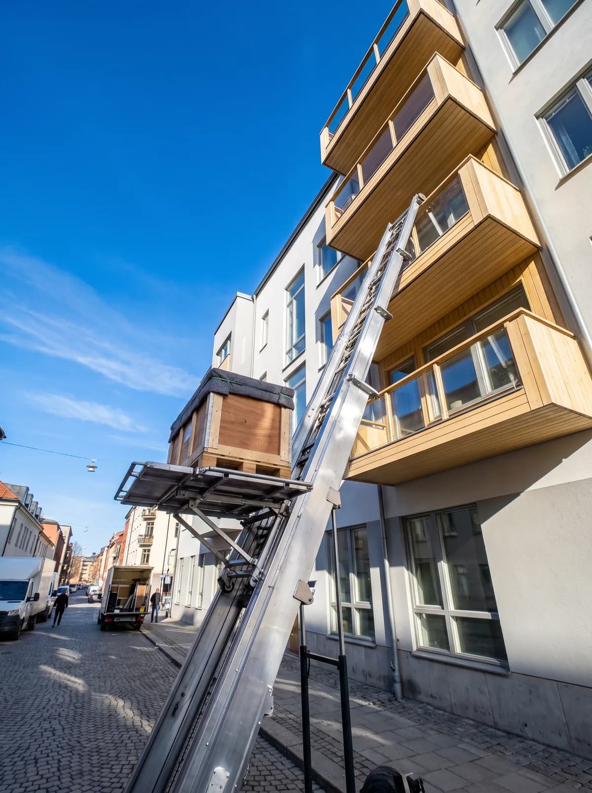 Möbelhiss som lyfter en flyttlåda upp till en balkong mot blå himmel